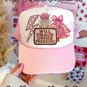 Puede incluir: Gorra de camionero rosa con un panel frontal blanco con un cascanueces de dibujos animados, un rayo rosa y un árbol de Navidad rosa. El texto "WILL CRACK UNDER PRESSURE" se muestra en un parche marrón. La gorra tiene una visera rosa y respaldo de malla.