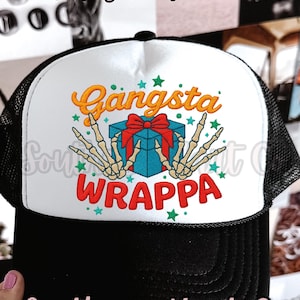 May include: Black and white trucker hat with a white front panel featuring the words "Gangsta WRAPPA" in orange and red, with a turquoise gift box and skeleton hands. The hat has a black brim and mesh back.