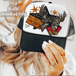 Pode incluir: Boné de caminhoneiro branco e preto com um design temático de caça. O boné apresenta uma estrela, um laço camuflado, chifres de veado e duas balas vermelhas. O texto "GIRLS HUNT TOO" é visível. Download digital.