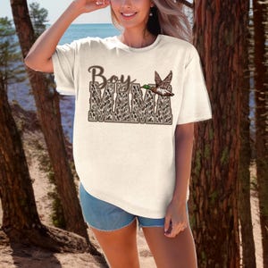 May include: A white t-shirt with a brown and white camouflage print that says "Boy Mama" with a duck flying above the text.