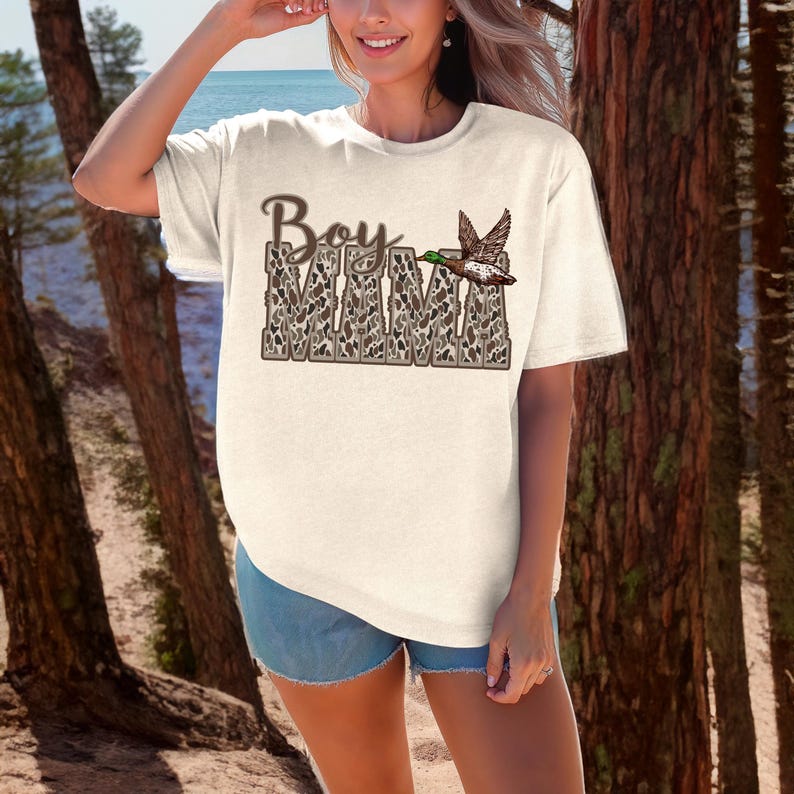 May include: A white t-shirt with a brown and white camouflage print that says "Boy Mama" with a duck flying above the text.