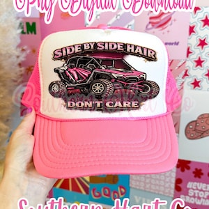 May include: Pink trucker hat with a white front panel featuring a pink off-road vehicle graphic and the text "SIDE BY SIDE HAIR DON'T CARE." The hat has a pink brim and mesh back. The text "Southern Hart Co" is also visible.