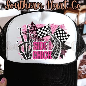 May include: Black and white trucker hat with a white front panel featuring a pink and black graphic. The graphic includes a skeleton hand, checkered flags, and the words "SIDE CHICK". The hat has a black brim and mesh back.