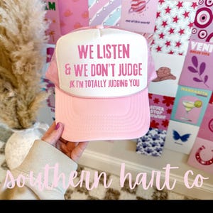 May include: Pink trucker hat with white mesh back. The hat has pink text that reads "We listen & we don't judge JK I'm totally judging you".