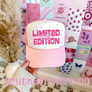 May include: Pink trucker hat with a white mesh back. The front of the hat has a white background with pink, blue, and yellow stars and the text "Limited Edition" in pink with a pink heart.