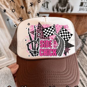 May include: A brown and white trucker hat with a brown peak and mesh back. The front panel has a pink and black design with the words "Side Chick", chequered flags, and a skeleton hand. The text "Southern Hart Co" is at the bottom.