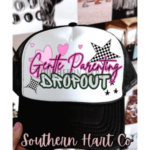 May include: Black and white trucker hat with a white front panel. The hat features the text "Gentle Parenting Dropout" in pink and gray, along with pink hearts, stars, and checkered flag designs.