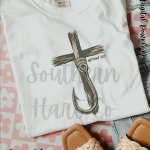 K&ouml;nnte beinhalten: Ein wei&szlig;es kurz&auml;rmeliges T-Shirt mit einer Grafik eines dunkelbraunen Kreuzes, das mit einem Angelhaken verschlungen ist. Der Text Matthew 4:19 ist neben dem Kreuz aufgedruckt. Das T-Shirt hat ein Comfort Colors Etikett am Innenkragen.