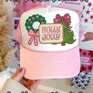 May include: A pink trucker hat with a white front panel. The panel displays a Christmas wreath, a Christmas tree, and the words "HOLLY JOLLY". The hat has a pink brim and mesh backing. The text "Png Digital Download" and "Southern Hart Co" are also visible.