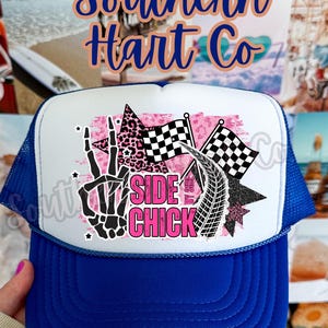 May include: A blue and white trucker hat with the text "Side Chick". The graphic features a skeleton hand, chequered flags, and a tyre track design. The hat has a blue brim and a mesh back.