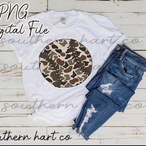 May include: A white t-shirt with a camouflage baseball graphic. The shirt is laying on a wooden surface with a pair of blue jeans.