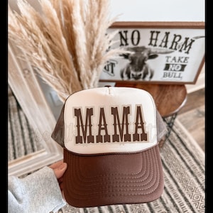 May include: A brown and white trucker hat with the word "MAMA" in brown letters. The hat has a brown brim and a white front panel. The image also shows a sign that says "NO HARM TAKE NO BULL".