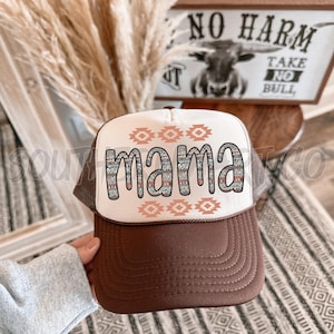 Peut inclure: Un chapeau de camionneur brun et blanc avec un panneau avant blanc orné d'un motif géométrique coloré et du mot "mama" dans une police de caractères colorée. Le chapeau a un dos en maille brune.