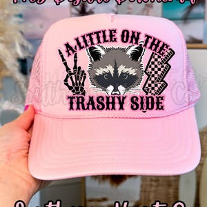 May include: Pink trucker hat with a mesh back and a curved brim. The front of the hat features a raccoon graphic and the text "A LITTLE ON THE TRASHY SIDE". The hat also has a peace sign and checkered flag design.