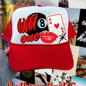 Puede incluir: Gorra de camionero roja y blanca con las palabras "Wild One" en escritura roja, una bola 8, una carta de juego y labios rojos. La gorra tiene una visera roja y una parte trasera de malla.