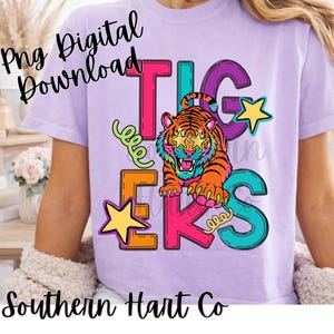 May include: A light purple t-shirt featuring a graphic design with the word TIGERS in colorful block letters, a tiger with star-shaped glasses, and yellow stars. The text Png Digital Download and Southern Hart Co are visible on the image.