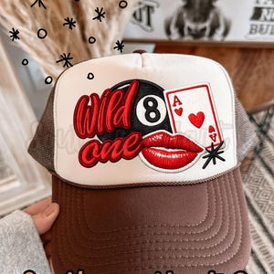 Puede incluir: Gorra de camionero marrón y blanca con la parte trasera de malla marrón. El panel frontal presenta las palabras "Wild One" en escritura roja, una bola 8 negra, una carta de juego con un corazón y labios rojos. El texto "Southern Hart Co" está en la parte inferior.