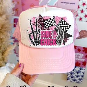 May include: Pink trucker hat with a white front panel featuring a graphic that includes a skeleton hand, checkered flags, and the words "SIDE CHICK" in pink. The hat has a pink brim and mesh back.