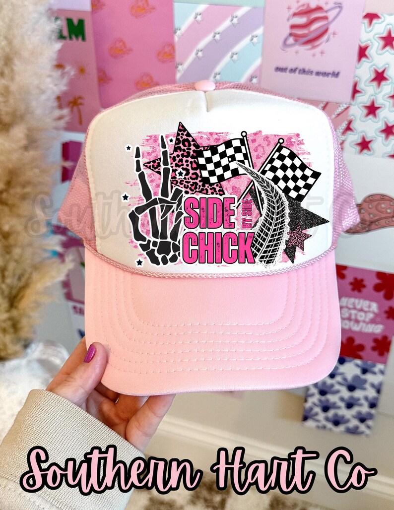 May include: Pink trucker hat with a white front panel featuring a graphic that includes a skeleton hand, checkered flags, and the words "SIDE CHICK" in pink. The hat has a pink brim and mesh back.