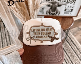 May include: A brown and white trucker hat with the text "Living on that Blue Collar Dollar" and a steer head graphic. The hat has a brown brim and mesh back, with the words "No Harm Take No Bull" in the background.