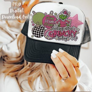 May include: A black and white trucker hat with a white front panel featuring a pink and green Grinch design with the text "STINK STANK STUN" and "feeling extra GRINCHY today".