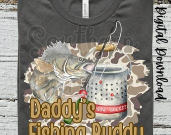 Pode incluir: Camiseta cinza escuro com um peixe, uma vara de pesca e um balde com o texto "Daddy's Fishing Buddy". O design inclui um padrão de camuflagem e as palavras "Southern Hart Co."