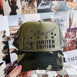 Peut inclure: Casquette de camionneur verte avec une visière camouflage et un dos en filet. L'avant présente un crâne de longhorn, des cactus, des étoiles et les mots "LIMITED EDITION". Le texte "Southern Hart Co" est également visible.