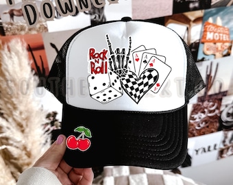 Pode incluir: Boné de caminhoneiro preto e branco com parte traseira em malha e aba curva. O painel frontal apresenta um design "Rock n Roll" com dados, cartas de baralho e um coração quadriculado. Um emblema de cereja está na aba. O texto "PNG DIGITAL DOWNLOAD" e "SOUTHERN HART CO" também são visíveis.