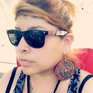 May include: A woman wearing black sunglasses with a colorful design on the frame and large, round, colorful earrings. She is wearing a black tank top with a blue and black patterned scarf.