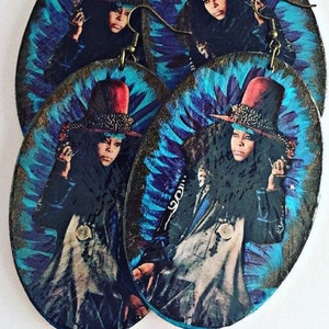 May include: A pair of wooden earrings featuring a colourful image of a woman wearing a red hat and a black jacket. The earrings have a blue and purple tie-dye background.