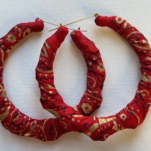 May include: A pair of large hoop earrings made from red fabric with a gold paisley pattern. The fabric is twisted and wrapped to create a unique and textured look.