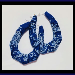 May include: A pair of blue bandana hoop earrings. The earrings are made of fabric with a white paisley pattern. The hoops are attached to silver-colored posts with clear earring backs. The earrings are displayed on a white surface.