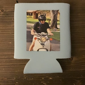 May include: A blue can cooler with a photo of a person wearing a helmet and riding a white scooter.