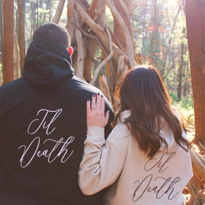 May include: A couple wearing matching black and beige hoodies with the text "Til Death" written in white cursive on the back.