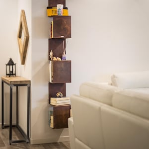May include: A white couch with pillows sits in front of a wall with a vertical shelf unit. The shelf unit is made of metal and has multiple shelves. The shelves are filled with books, decorative items, and a small plant. The shelf unit is painted in a rusty brown color.