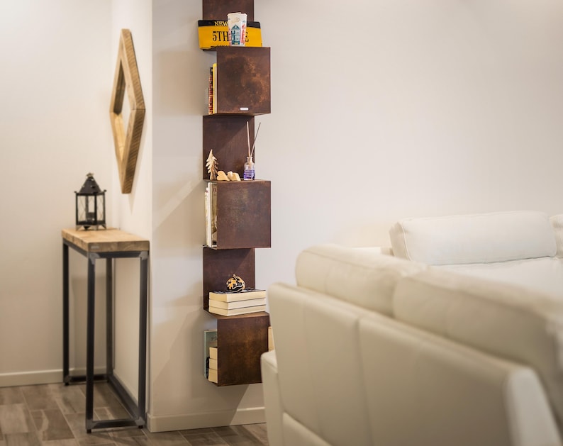 May include: A white couch with pillows sits in front of a wall with a vertical shelf unit. The shelf unit is made of metal and has multiple shelves. The shelves are filled with books, decorative items, and a small plant. The shelf unit is painted in a rusty brown color.