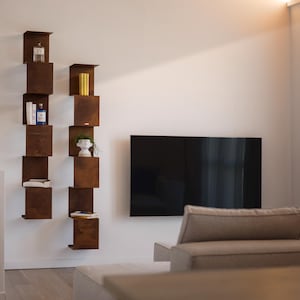 May include: Two vertical, staggered, open shelves made of brown metal with various items on the shelves, including books, a vase, and a small plant. A flat screen TV is mounted on the wall behind the shelves. A light brown couch is in the foreground.