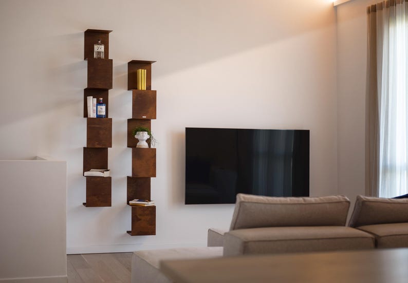May include: Two vertical, staggered, open shelves made of brown metal with various items on the shelves, including books, a vase, and a small plant. A flat screen TV is mounted on the wall behind the shelves. A light brown couch is in the foreground.