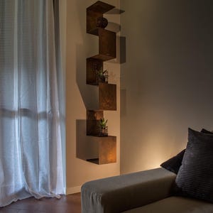 May include: A living room with a brown couch and a set of brown metal shelves with plants on them. The shelves are mounted on a wall with a light brown paint color. The couch has a light brown fabric cover and two pillows. The floor is a dark brown tile.