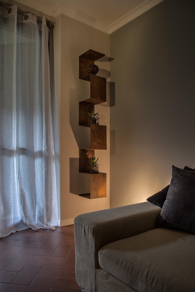 May include: A living room with a brown couch and a set of brown metal shelves with plants on them. The shelves are mounted on a wall with a light brown paint color. The couch has a light brown fabric cover and two pillows. The floor is a dark brown tile.