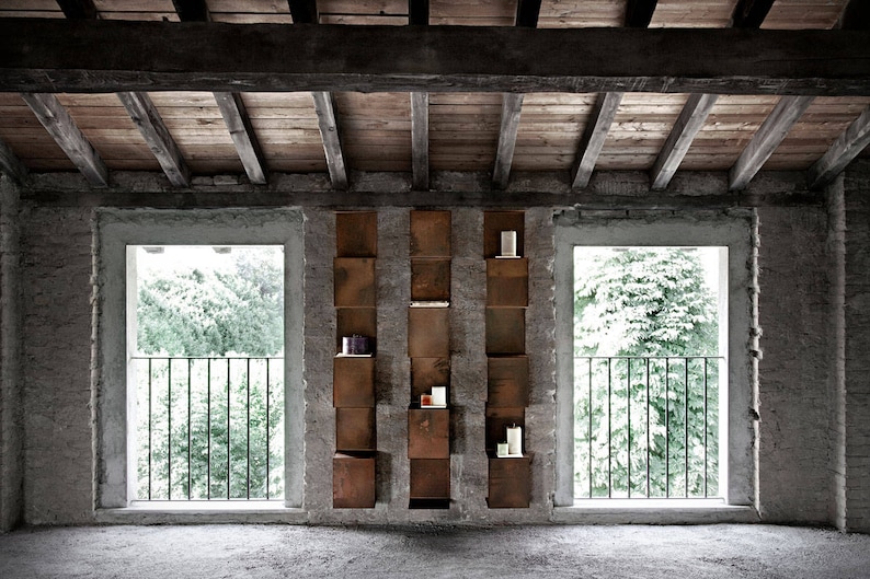 May include: A rustic interior with two windows overlooking a green tree-lined view. A wall features a built-in shelving unit with a geometric design, made of a dark, rusted metal.