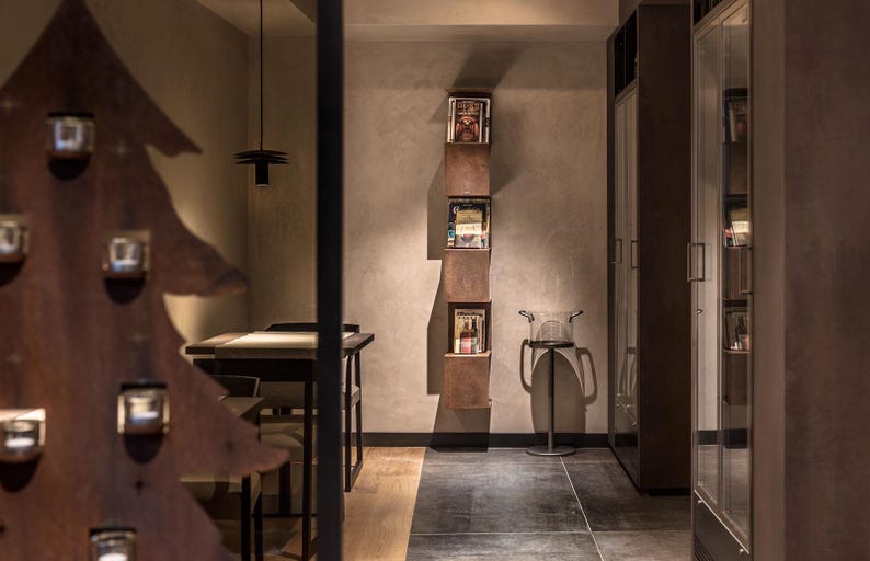 May include: A modern interior design with a brown wooden tree sculpture, a dark wood table with chairs, and a wall-mounted shelf with magazines. The shelf is made of metal and has a rustic look.