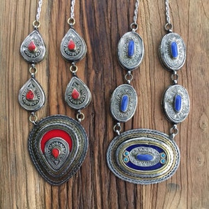 May include: Two silver necklaces with pendants. One necklace features red stones and a large teardrop-shaped pendant. The other necklace features blue stones and a large oval pendant.