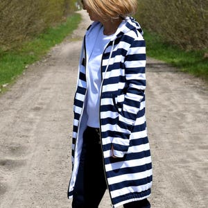 Könnte beinhalten: Eine lange, marineblau-weiß gestreifte Kapuzenjacke mit durchgehendem Reißverschluss. Die Jacke wird über einem weißen T-Shirt und einer dunkelblauen Hose getragen. Weiße High-Top-Sneaker vervollständigen das lässige Outfit.
