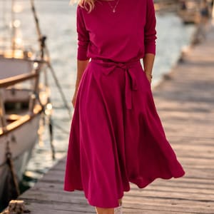 May include: A magenta dress with a long-sleeved top and a flowing skirt, tied at the waist with a matching belt. The dress is being worn by a person walking on a wooden dock, wearing white trainers. The background shows a body of water and a boat.