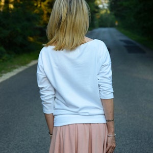 Puede incluir: Una mujer con una camisa blanca de manga larga y una falda plisada de color melocot&oacute;n. Est&aacute; caminando por un camino pavimentado.