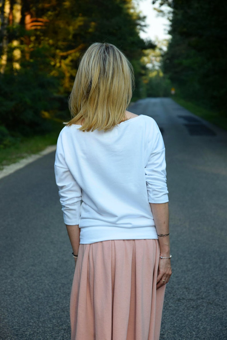 Puede incluir: Una mujer con una camisa blanca de manga larga y una falda plisada de color melocot&oacute;n. Est&aacute; caminando por un camino pavimentado.