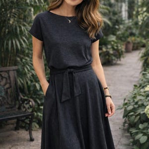 May include: A dark gray dress with short sleeves and a tie waist. The dress has a flared skirt and pockets. The model is wearing a necklace and bracelets. The background is a greenhouse with lush greenery.