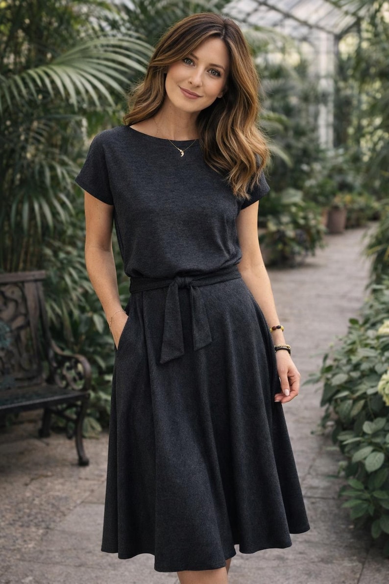 May include: A dark gray dress with short sleeves and a tie waist. The dress has a flared skirt and pockets. The model is wearing a necklace and bracelets. The background is a greenhouse with lush greenery.