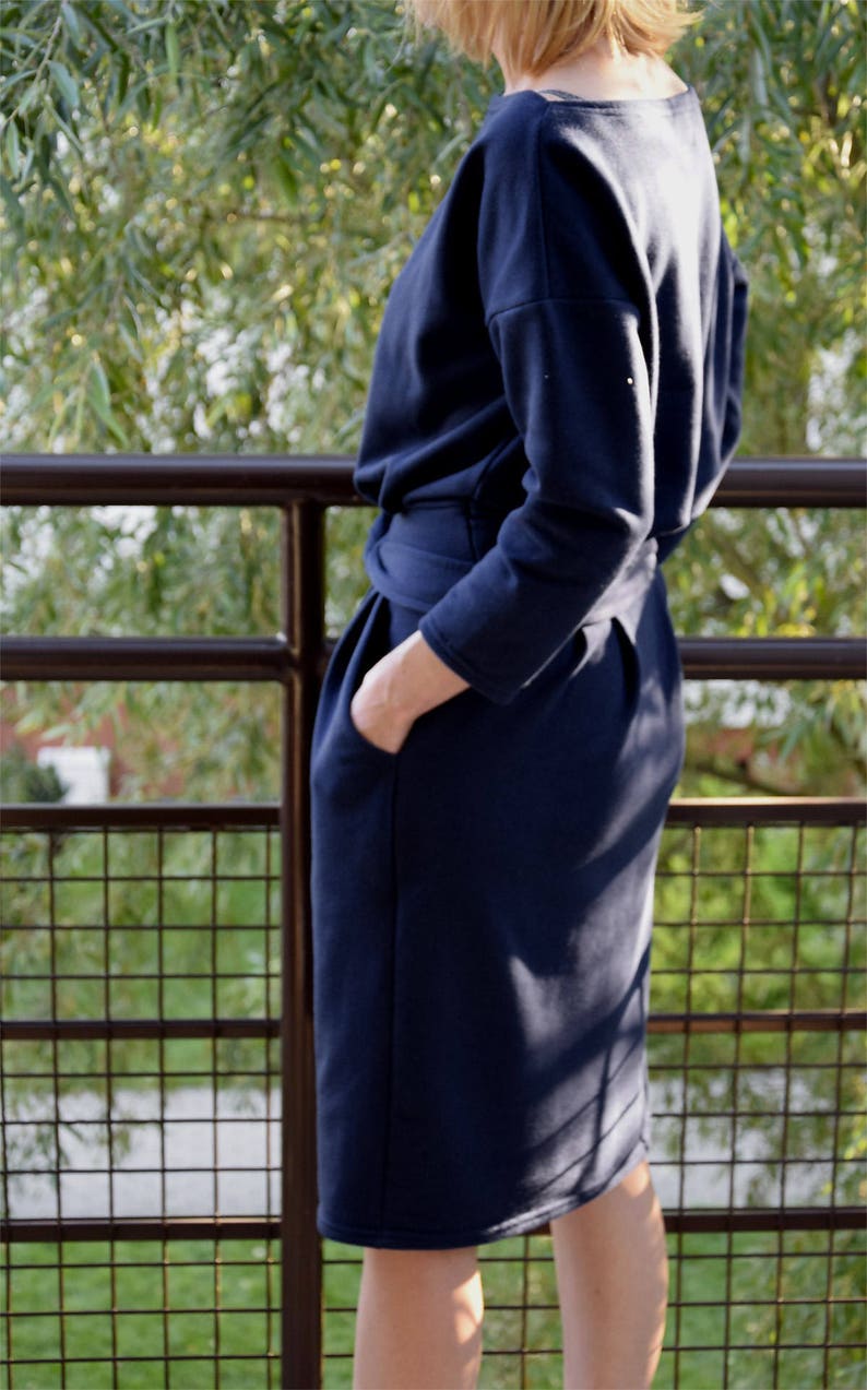 May include: A navy blue knee-length dress with long sleeves, a boat neckline, and a cinched waist featuring a tie belt. The garment includes side pockets and is constructed from a smooth, solid-colored fabric with a structured silhouette.